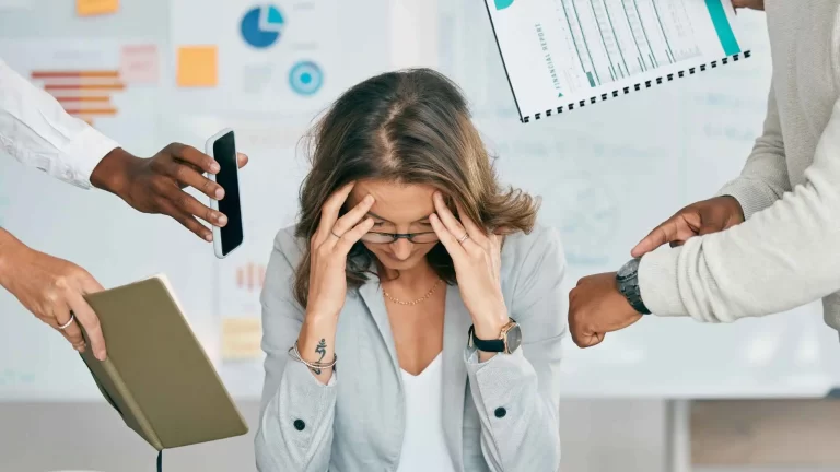 Mujer con las manos en la cabeza mientras diferentes personas le acercan documentos, celular y le muestran el relok prevenir el burnout con amor Magui Block
