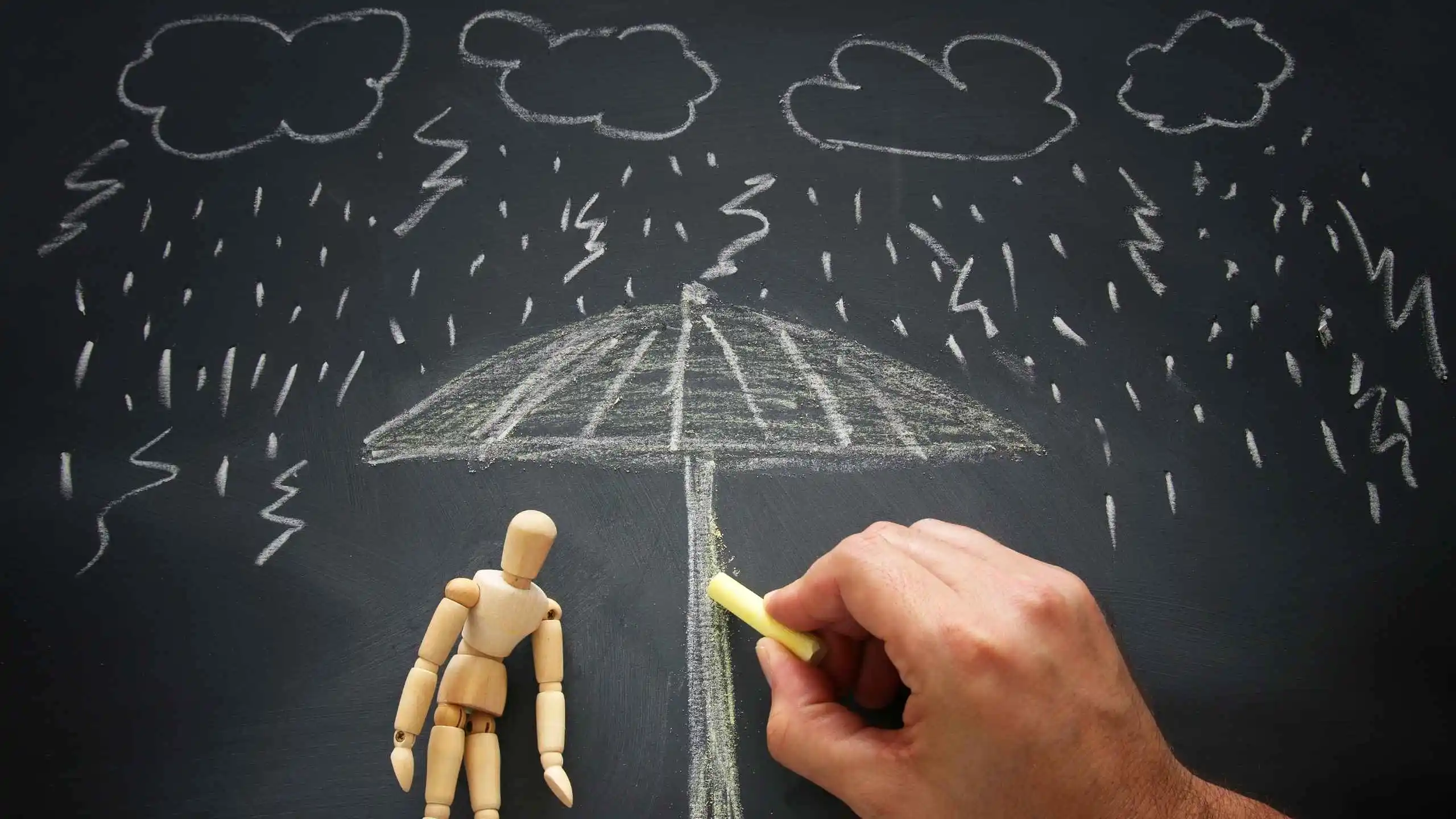 dibujo en tiza de un cielo nublado con rayos y un paraguas. Bajo el paraguas està un muñeco de madera y aparece también la mano que dibuja en el tablero qué es la ansiedad magui block
