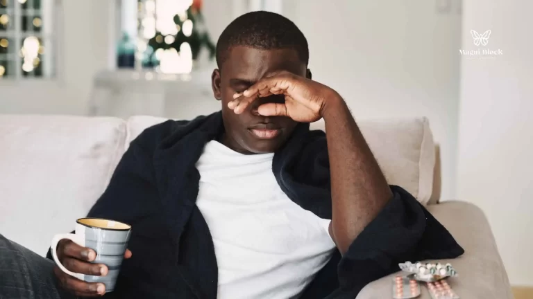 Hombre afrodescendiente con la mano sobre la cara y en la otra mano sostiene un pocillo sentado en un sofá beige y en el brazo del asiento hay medicamentos tipo píldora como afecta el cáncer la salud mental magui block