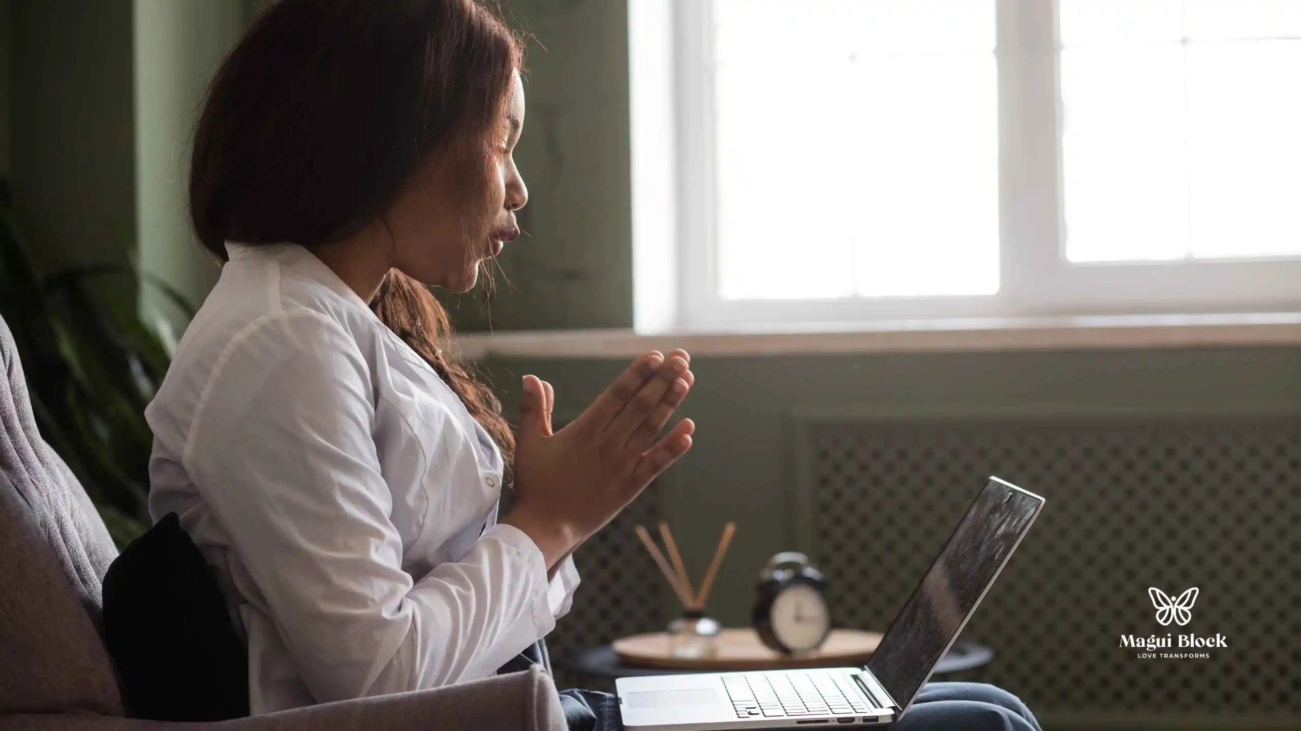 mujer afrodescendiente habla al computador que tiene en las piernas sentada en una silla individual al lado de una ventana terapia online efectiva magui block